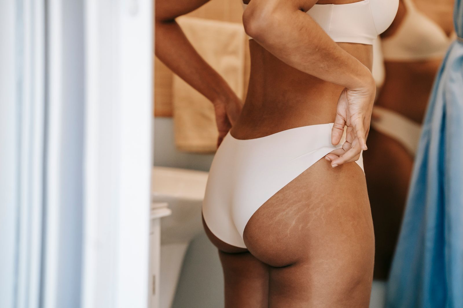Woman in cream underwear seen from behind, hand on lower back, stretch marks visible on hip and thigh