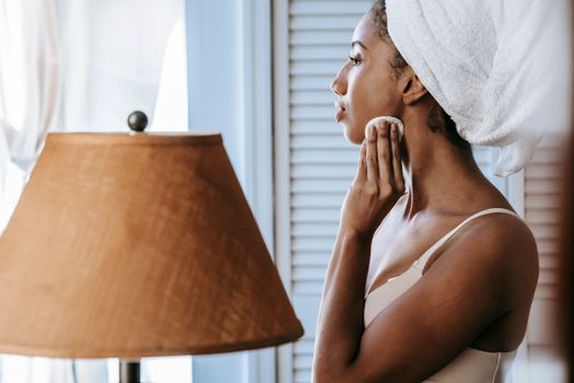 Side view of crop young ethnic female in brassiere cleaning face with cotton sponge at home while looking forward