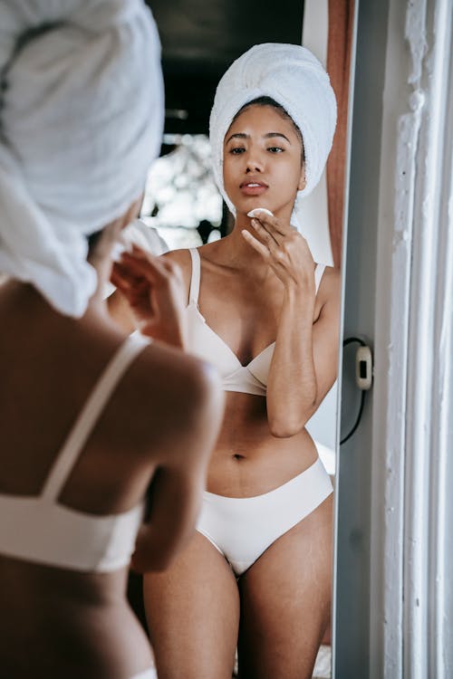 Ethnic woman in lingerie cleaning face against mirror at home