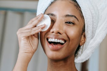 Cheerful ethnic female wrapped in towel turban moisturizing face with lotion while smiling happily