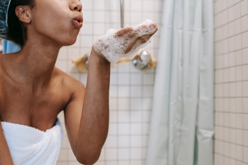 Side view of crop anonymous African American female in towel blowing off foam from hand in bathroom
