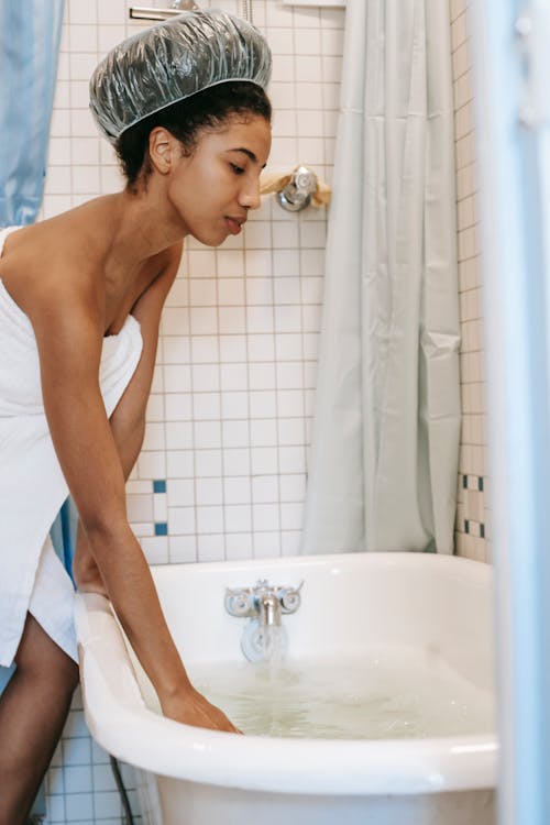 Side view of young African American female in white towel and shower cap standing near bathtub and touching water before washing
