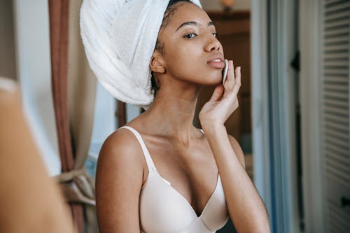 Young concentrated African American female in underwear with white towel on head wiping face with cotton pad