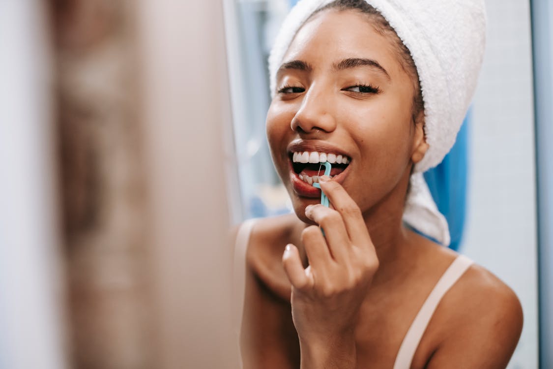 Smiling African American female with white towel on head cleaning teeth with dental flosser in bathroom