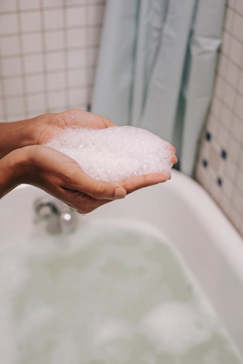 Crop anonymous female with bubbling foam in hands over bathtub while washing in bathroom