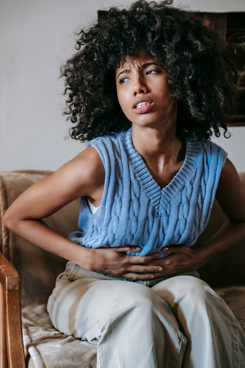 Free Young African American female sitting on couch at home while having acute pain in stomach Stock Photo
