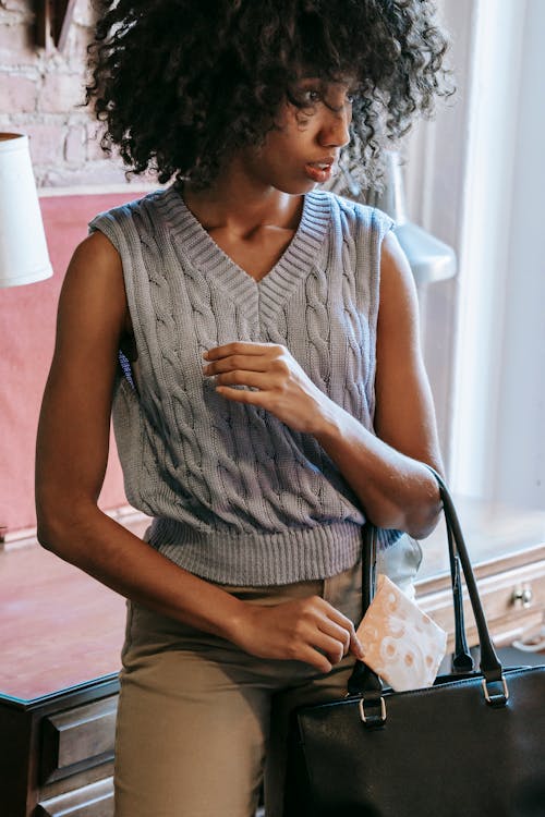 Young African American female in casual outfit putting sanitary pad in black leather handbag