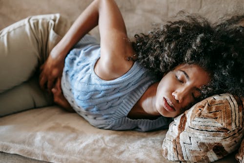 Free High angle of African American woman with afro hairstyle and closed eyes having pain in belly lying on sofa Stock Photo