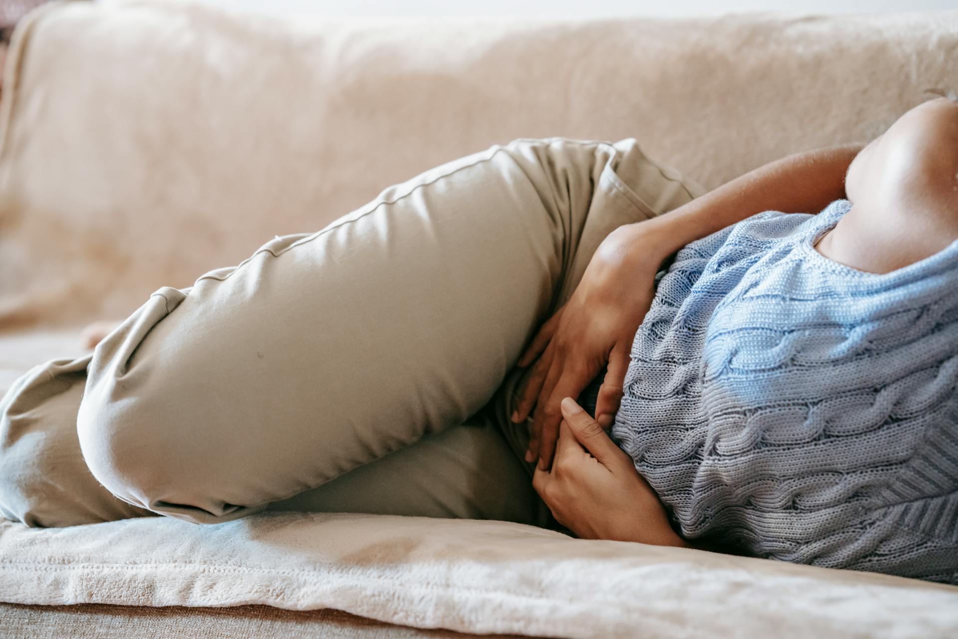 Crop faceless female in casual wear lying on sofa while having acute pain in stomach