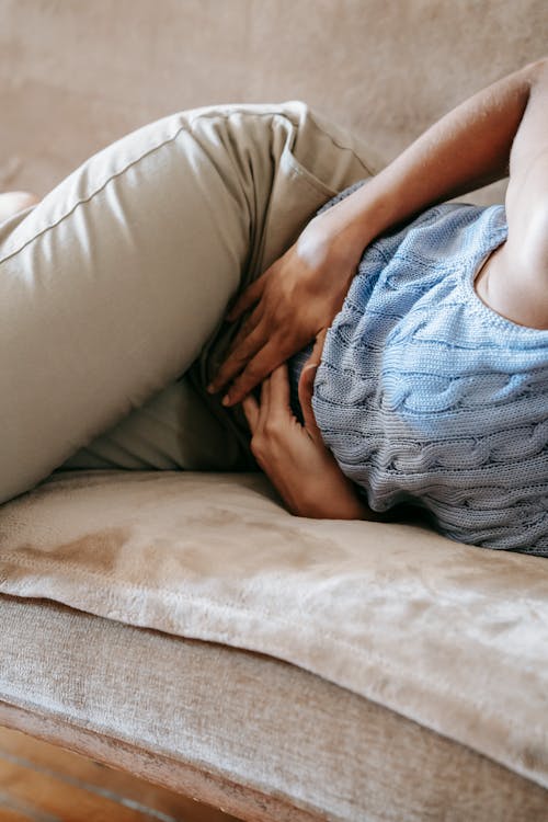 Crop anonymous female in casual outfit writhing on sofa while having stomach ache