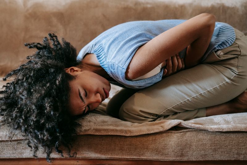 A Black woman curled on a couch holding her abdomen in pain, the kind of persistent upper-stomach burning that an H. pylori infection or peptic ulcer can cause.