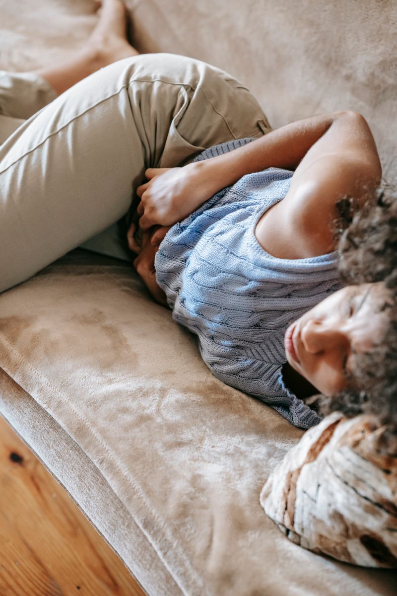 A Black woman lies on a couch holding her abdomen in pain, a scene that mirrors how acute pancreatitis often presents: severe upper-belly pain that can radiate to the back and worsen after eating.