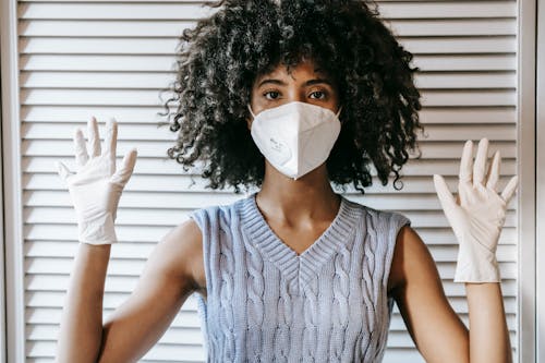 Young African American female wearing white protective face mask and medical white rubber gloves with hands raised looking at camera