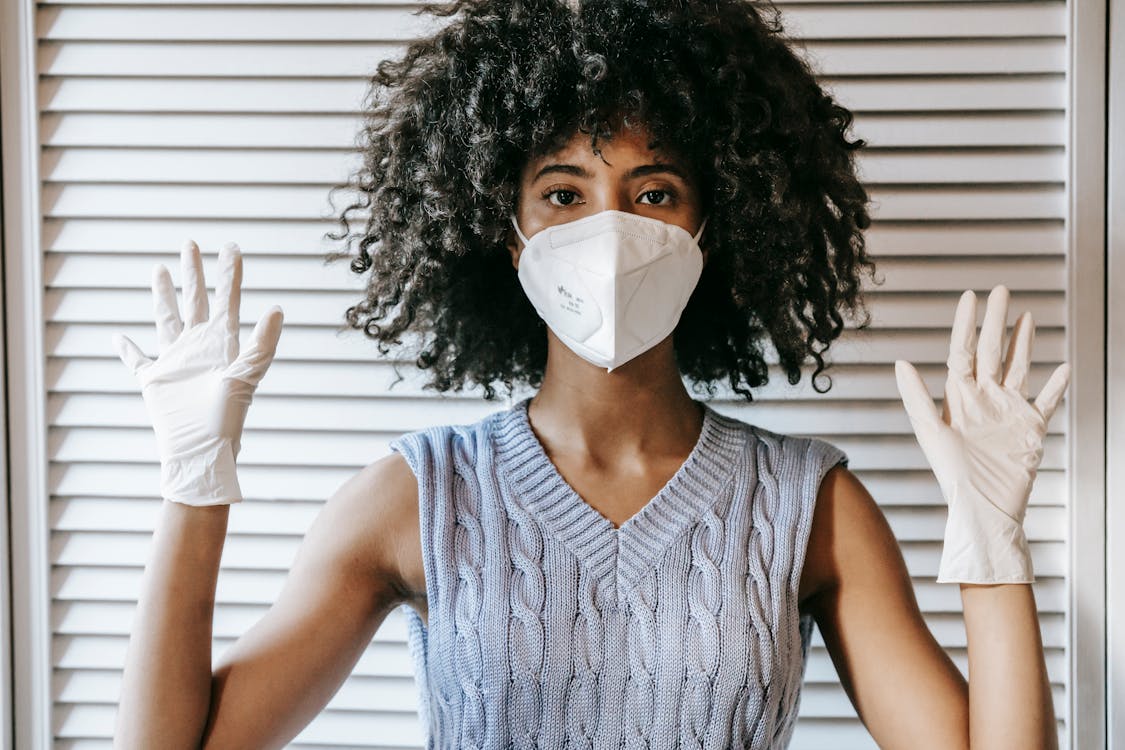 Free Young African American female wearing white protective face mask and medical white rubber gloves with hands raised looking at camera Stock Photo