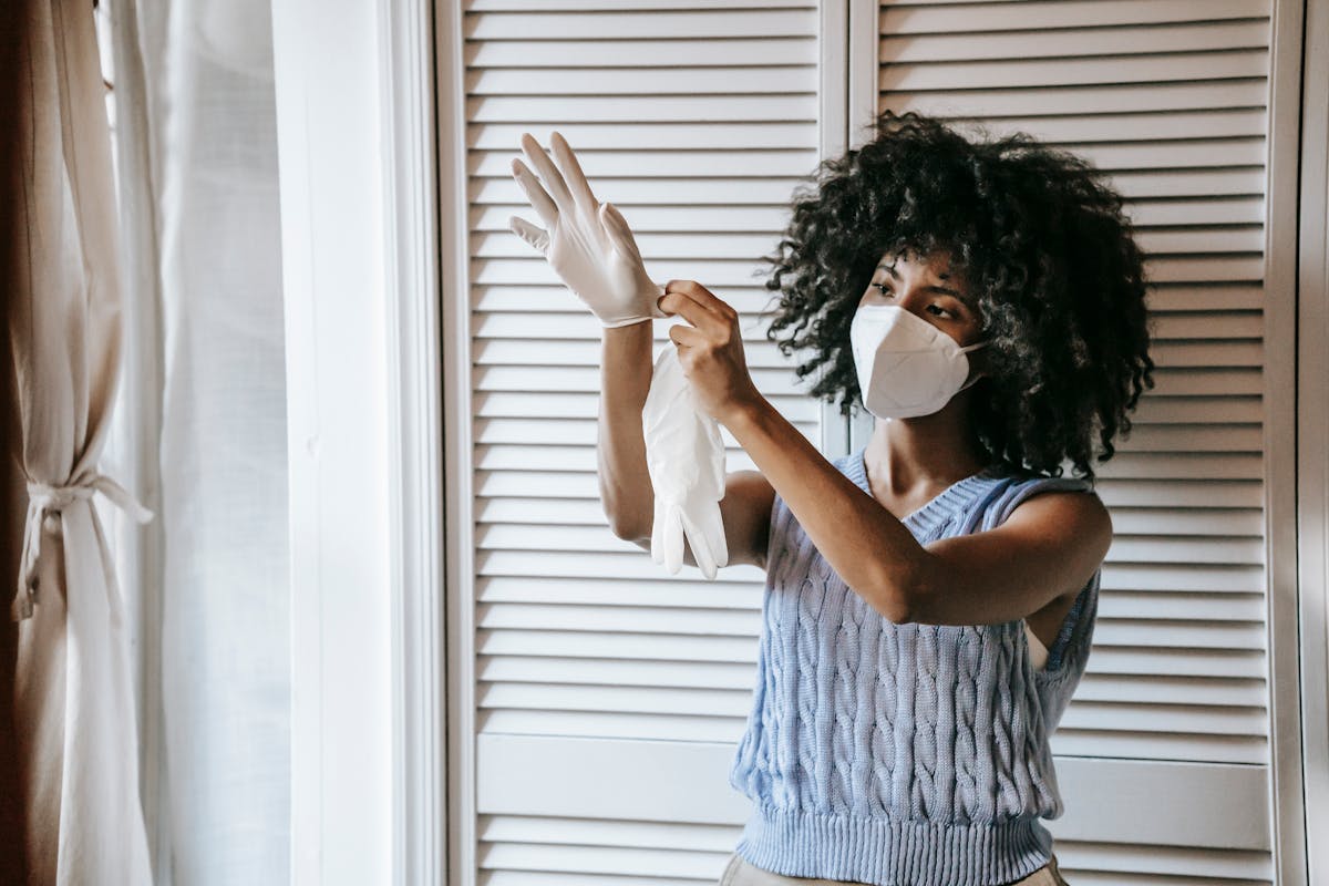 Young African American female in casual outfit and protective face mask putting off medical rubber gloves