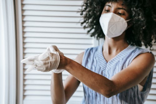 Young African American female in white protective face mask putting of medical latex gloves