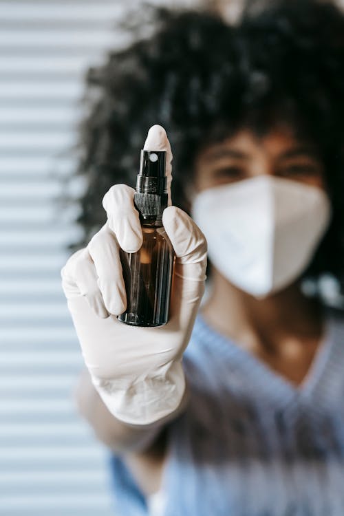 Blurred African American female wearing white protective mask and latex medical sterile gloves spraying antiseptic in room and looking at camera