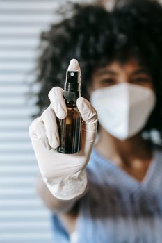 一名模糊的非裔美国女性戴着白色防护口罩和乳胶医用无菌手套，在房间里喷洒消毒剂并看着镜头