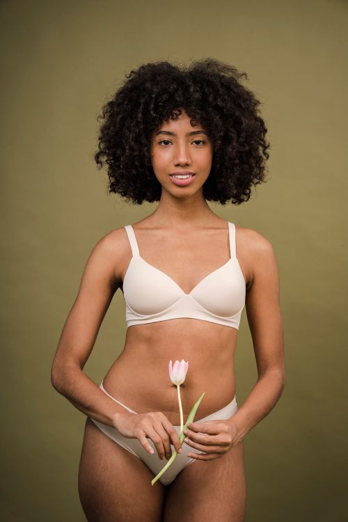 Free Graceful young African American lady with curly hair in white lingerie smiling in beige studio with delicate tulip flower in hands Stock Photo