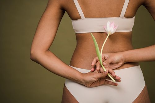 Free Back view of crop anonymous young female in white bra and panties holding fresh delicate tulip flower against beige background Stock Photo