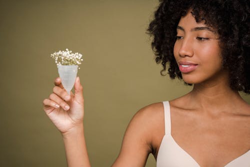 Happy young African American female model with curly hair in bra smiling and showing menstrual cup with gentle flowers against beige background