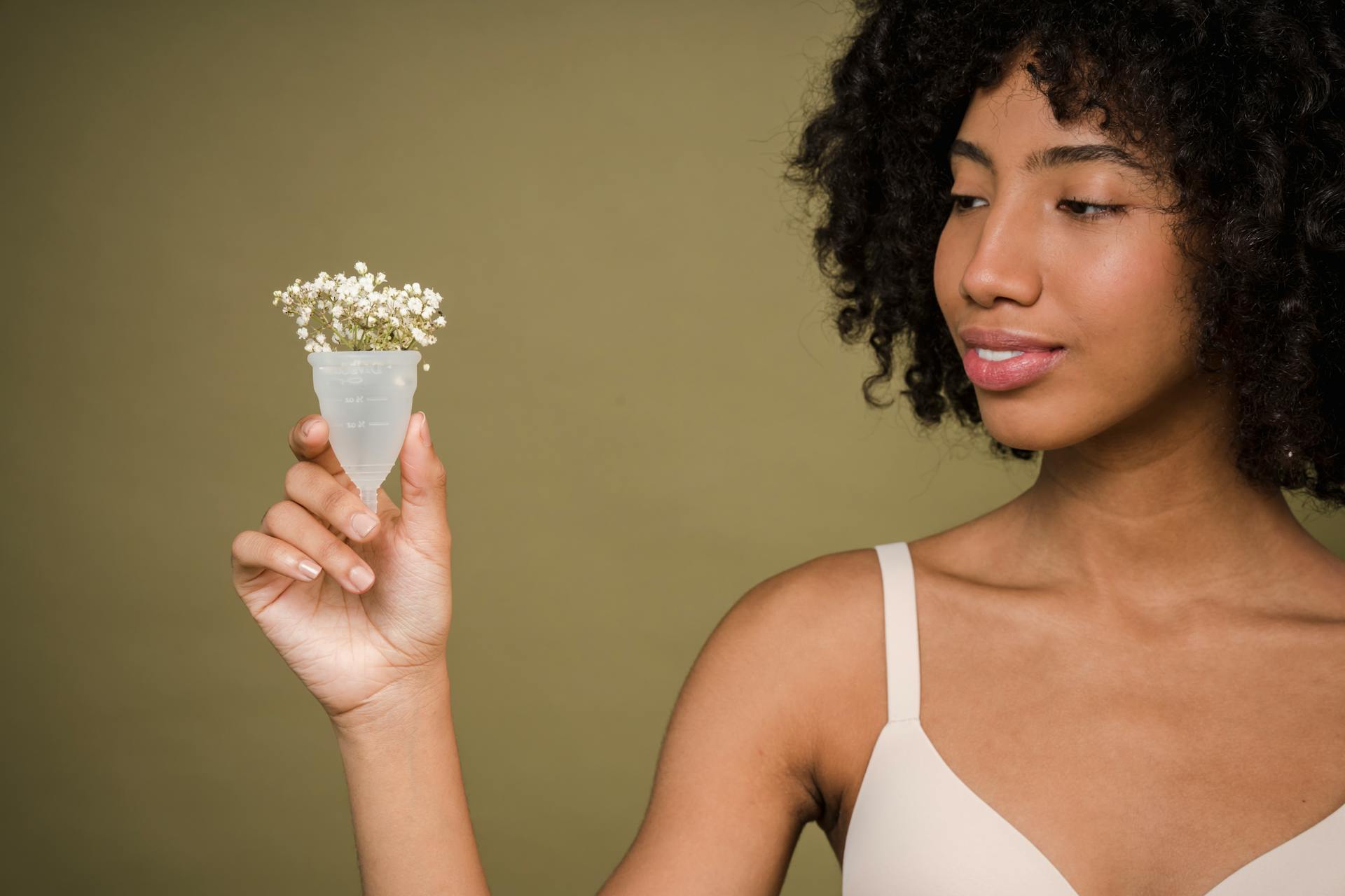 Happy young African American female model with curly hair in bra smiling and showing menstrual cup with gentle flowers against beige background