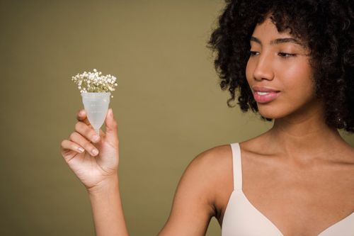 Free Happy young African American female model with curly hair in bra smiling and showing menstrual cup with gentle flowers against beige background Stock Photo