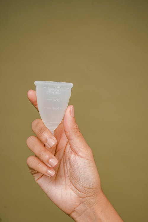 Crop faceless woman demonstrating small white silicone menstrual cup in hand against beige background