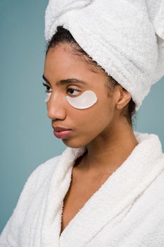 Pensive African American female with dark hair and eye patches under eyes in white bathrobe and towel on head looking away against blue background