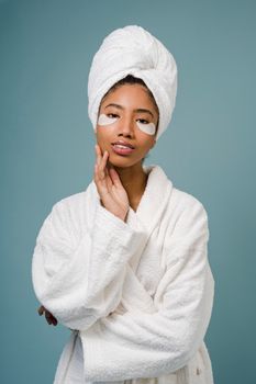 Cheerful African American woman with eye patches in white towel on head and bathrobe touching face and looking at camera against blue background