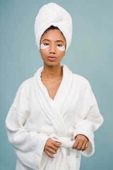 Calm African American female with moisturizer eye patches in white towel on head and bathrobe looking at camera against blue background in studio