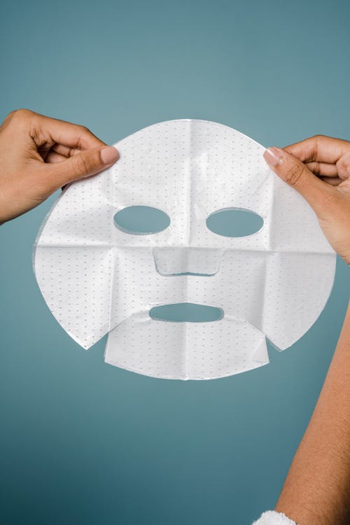 Crop unrecognizable ethnic woman demonstrating moisturizing sheet mask against blue background in studio