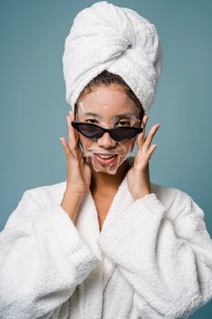 Cheerful black woman in stylish sunglasses and towel on head applying sheet mask and looking at camera against blue background