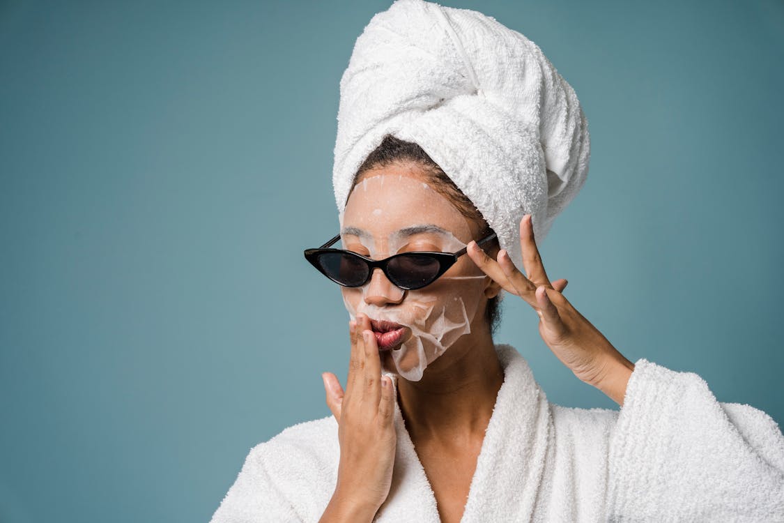 Calm African American woman with moisturizing sheet mask in sunglasses and white towel turban on head touching dace against blue background in studio