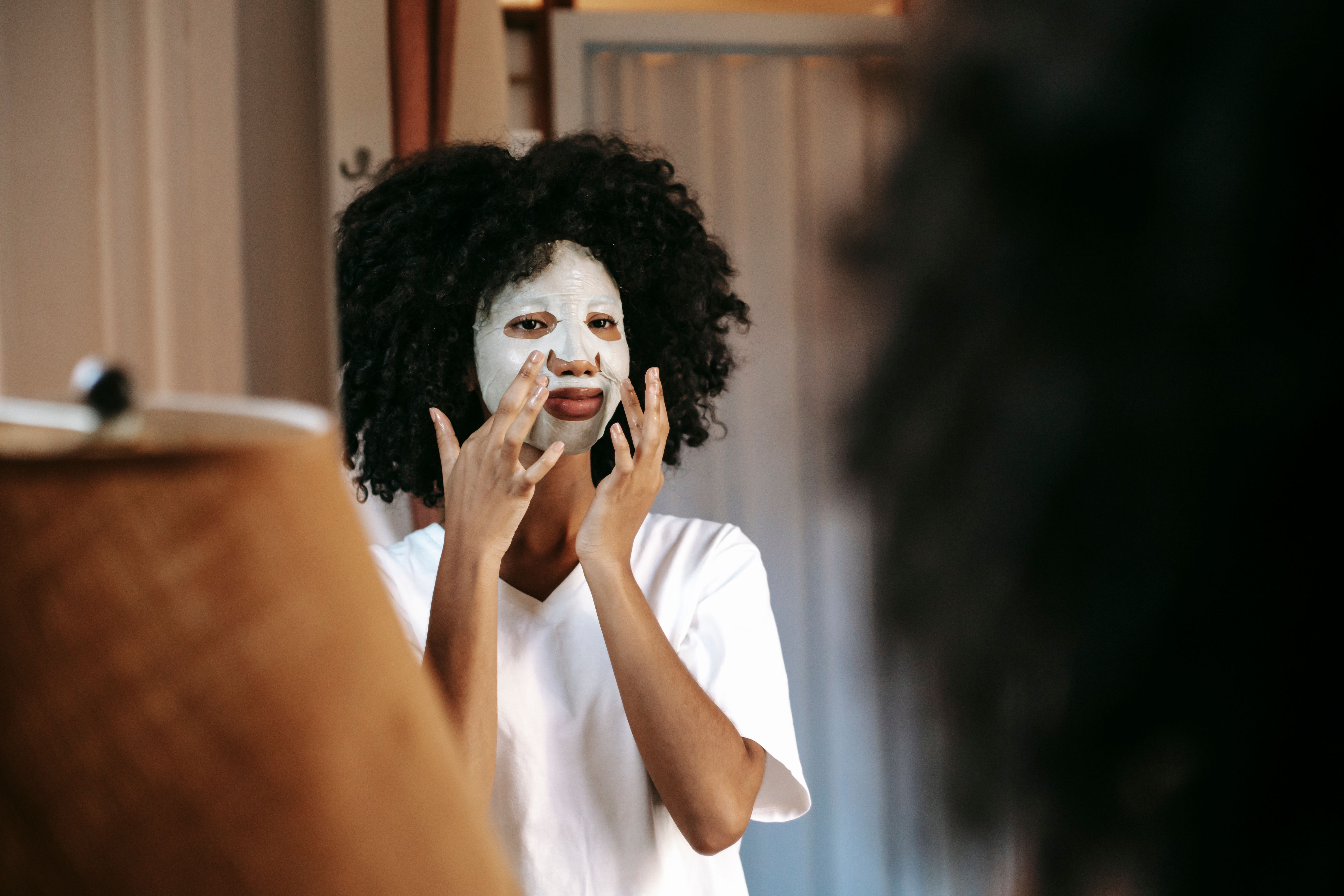 African American woman with dark curly hair applying sheet mask and looking at mirror in apartment in daytime