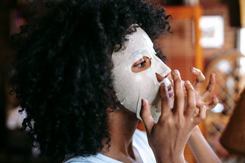 Side view of cheerful African American woman with black curly hair applying sheet mask at home and looking away in daylight