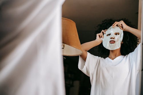 From below of black female with curly hair in white t shirt standing near mirror in room and applying sheet mask in daytime