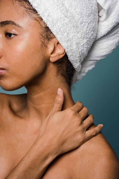 Side view of crop African American female in white towel on head looking away and touching naked shoulders against blue background