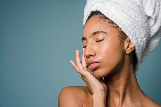 Charming naked black female with towel turban on head standing with closed eyes and touching face against blue background in studio