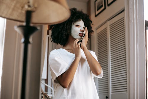 From below of African American female with dark curly hair with sheet mask in casual clothes standing in room and touching face in daytime