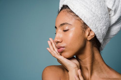 Charming African American female with towel on head and bare shoulders touching face with closed eyes after shower against blue background in studio