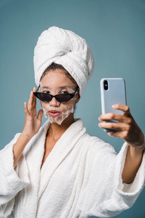 Confident black female with sheet mask taking selfie on smartphone