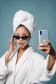 Stylish woman in bathrobe and sheet mask taking selfie on blue background.