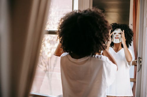 Back view of calm African American woman in casual clothes applying sheet mask and looking at mirror in light room at home in daytime