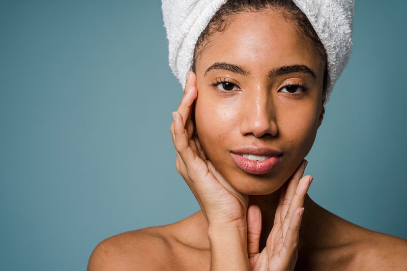 A Black woman with a towel wrapped around her hair rests her hands against her cheeks, her bare face turned to the camera.