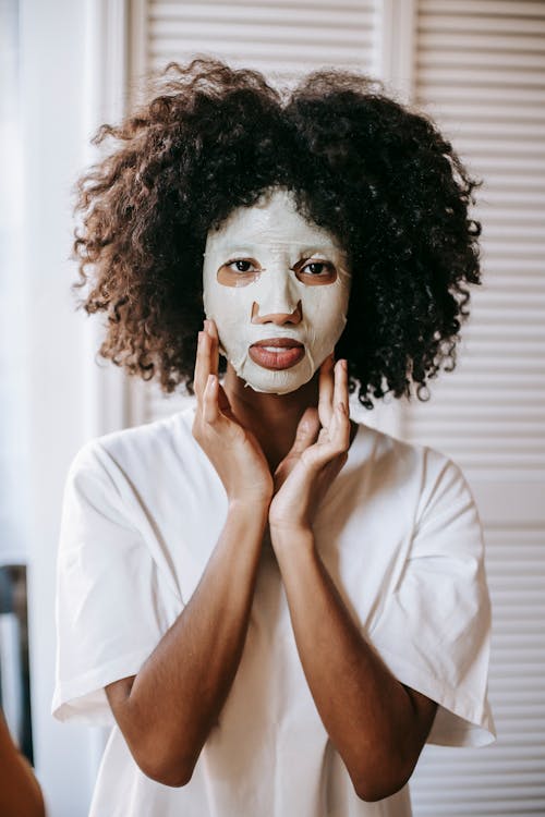 Pensive African American woman in sheet mask touching face and looking at camera at home in daytime