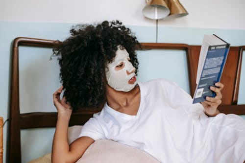 African American female with dark curly hair in sheet mask and casual clothes lying on bed and reading book at home in daytime
