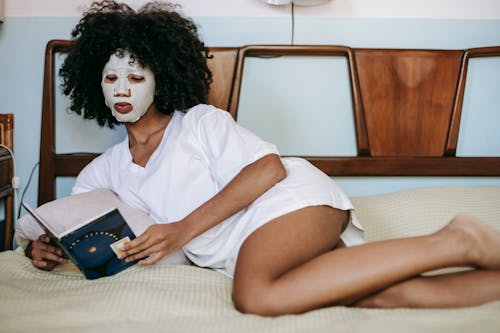 Young ethnic female in white t shirt relaxing on bed and reading book while doing facial routine with cosmetic sheet mask on face