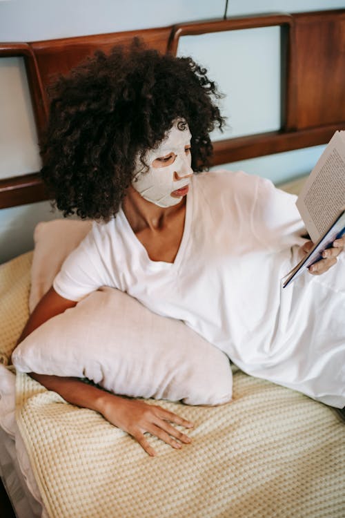 High angle of young ethnic female with cosmetic sheet mask on face lying on bed and reading book in bedroom