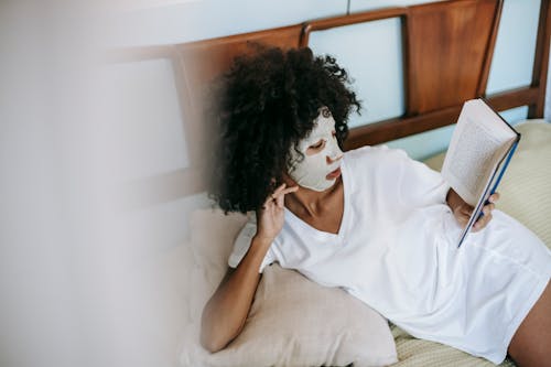 Young ethnic female in white t shirt with facial cosmetic mask on face having rest on bed and reading book